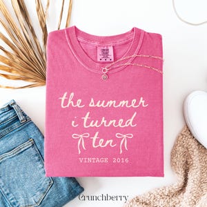 May include: A bright pink short-sleeve t-shirt featuring the white text "the summer i turned ten" with two bow illustrations and the text "VINTAGE 2016" below. The shirt has a crew neckline and a Comfort Colors brand label at the inner collar.