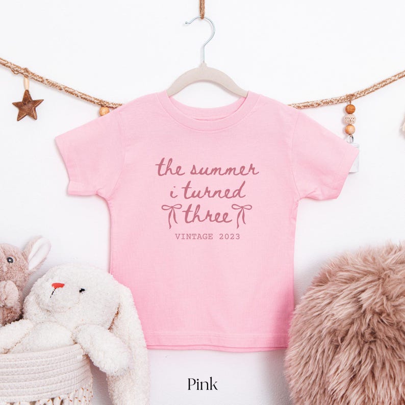 Puede incluir: Una camiseta rosa con el texto "the summer i turned three" y "VINTAGE 2023" en cursiva. La camiseta est&aacute; colgada de una percha. Un conejito de peluche y una cesta est&aacute;n en la esquina inferior izquierda. La palabra "Pink" est&aacute; abajo.