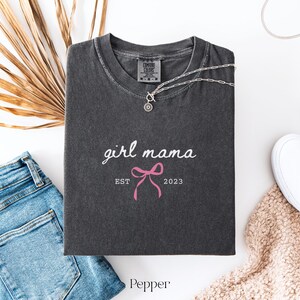 May include: A charcoal gray short-sleeve t-shirt with a crew neckline. The shirt features the white text girl mama, a pink bow graphic, and the white text EST 2023. The inner neck label displays the Comfort Colors brand logo and size M.