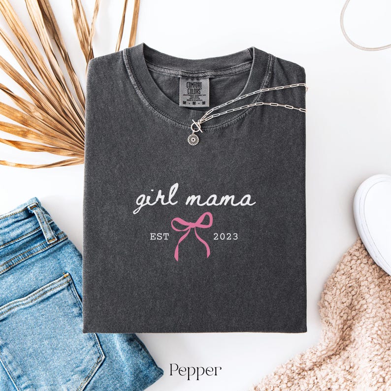 May include: A charcoal gray short-sleeve t-shirt with a crew neckline. The shirt features the white text girl mama, a pink bow graphic, and the white text EST 2023. The inner neck label displays the Comfort Colors brand logo and size M.