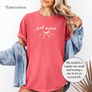 May include: A watermelon-colored t-shirt with the text "girl mama EST 2022" in white script. The shirt has a relaxed fit and is worn with a denim jacket. The model is wearing a size XL for an oversized fit.