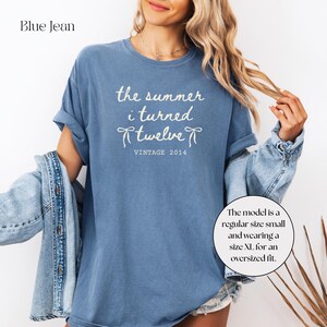 May include: Blue jean-colored t-shirt with the text "the summer i turned twelve VINTAGE 2014" in white. The shirt has short sleeves and a relaxed fit. The model is wearing a size XL for an oversized fit.