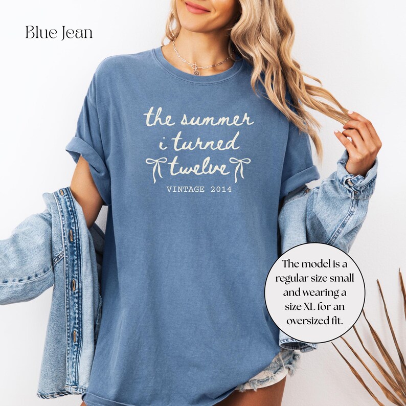May include: Blue jean-colored t-shirt with the text "the summer i turned twelve VINTAGE 2014" in white. The shirt has short sleeves and a relaxed fit. The model is wearing a size XL for an oversized fit.