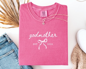May include: A pink short-sleeved t-shirt featuring the white text godmother, a graphic of a bow, and the text EST 2026. The shirt has a Comfort Colors brand label at the inner neckline and is displayed with a gold-toned chain necklace.