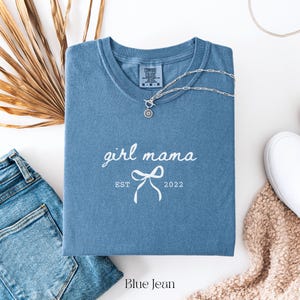 May include: A blue jean-colored t-shirt with the words "girl mama" and a bow graphic printed in white. The text "EST 2022" is below the bow. A silver necklace with a pendant is on the shirt. A pair of blue jeans and a white shoe are also in the image.