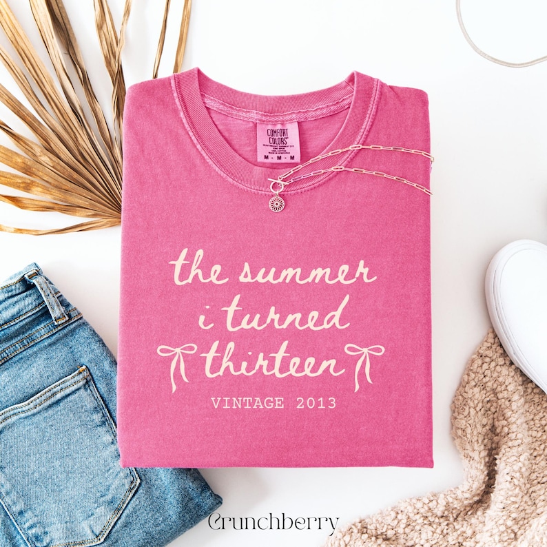Pu&ograve; includere: Una maglietta Comfort Colors rosa con la scritta "the summer i turned thirteen" e "Vintage 2013" in corsivo bianco. Una collana d'argento e jeans blu sono accanto alla maglietta.