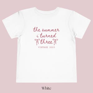 Puede incluir: Camiseta blanca para ni&ntilde;os peque&ntilde;os con el texto "the summer i turned three" en fuente cursiva, con dos gr&aacute;ficos de lazo y "VINTAGE 2023" debajo. La camiseta es de color blanco liso.