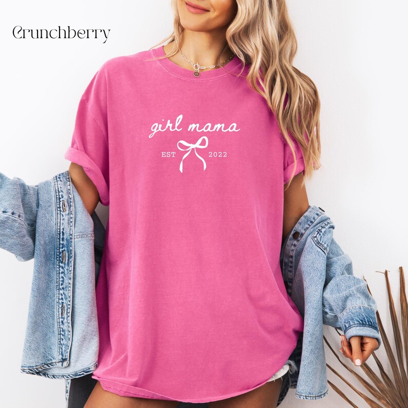 May include: A bright pink short-sleeved t-shirt featuring the white text girl mama, a small bow graphic, and the text EST 2022. The shirt is worn with a light blue denim jacket draped over the shoulders, showing a casual, loose-fitting style.