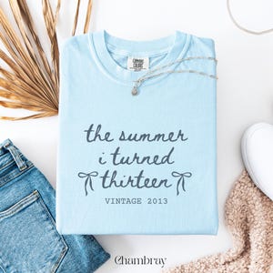 Pu&ograve; includere: Una maglietta azzurra con la scritta "the summer i turned thirteen" e "VINTAGE 2013". Una collana d'argento con un piccolo ciondolo &egrave; sulla maglietta. Chambray &egrave; stampato in basso. Jeans in denim e una sciarpa beige sono anche nell'immagine.