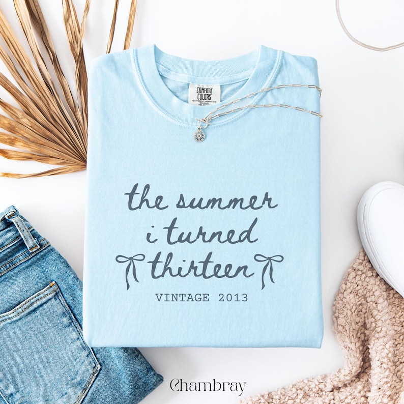Pu&ograve; includere: Una maglietta azzurra con la scritta "the summer i turned thirteen" e "VINTAGE 2013". Una collana d'argento con un piccolo ciondolo &egrave; sulla maglietta. Chambray &egrave; stampato in basso. Jeans in denim e una sciarpa beige sono anche nell'immagine.