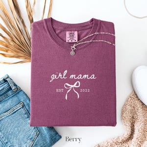 May include: A berry-colored t-shirt with the words "girl mama" and "EST 2022" printed in white. A silver necklace with a pendant is draped across the shirt. The shirt is folded, with a pair of blue jeans and a white shoe visible.