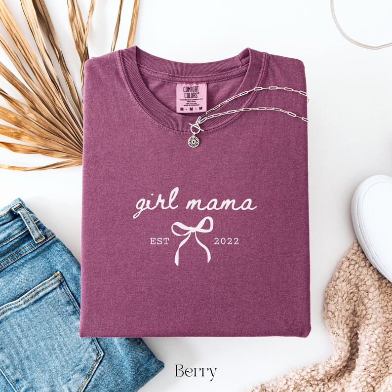May include: A berry-colored t-shirt with the words "girl mama" and "EST 2022" printed in white. A silver necklace with a pendant is draped across the shirt. The shirt is folded, with a pair of blue jeans and a white shoe visible.
