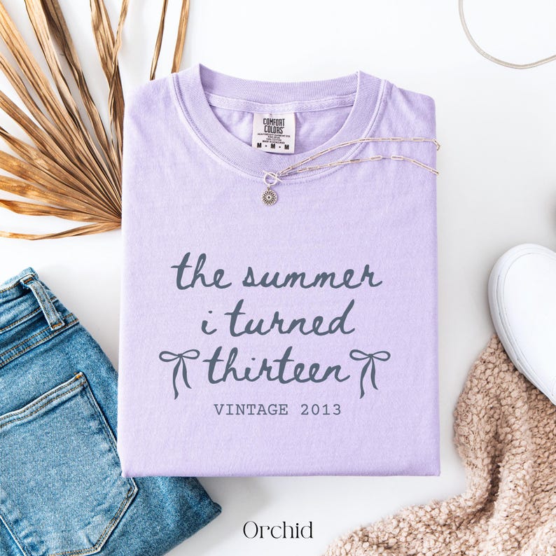 Pu&ograve; includere: Una maglietta Comfort Colors lilla con la scritta "the summer i turned thirteen" e "Vintage 2013". Una collana d'argento con un ciondolo a forma di sole &egrave; appoggiata sulla maglietta. La parola "Orchid" &egrave; in basso.
