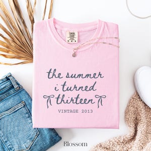 Pu&ograve; includere: T-shirt rosa chiaro con la scritta "the summer i turned thirteen" in grafia grigio scuro. La maglietta presenta anche la scritta "VINTAGE 2013" e la parola "Blossom". Una collana d'oro con un ciondolo &egrave; sulla maglietta.