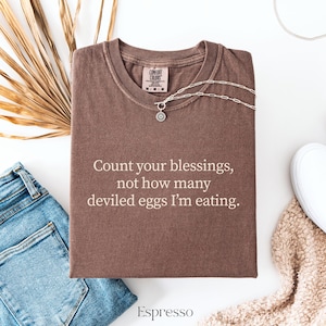 May include: A brown short-sleeve t-shirt featuring the text "Count your blessings, not how many deviled eggs I'm eating." printed in a light beige font. The shirt has a crew neckline and a Comfort Colors brand label on the inner collar.