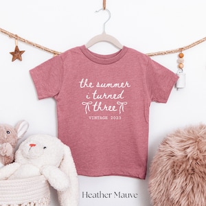 Puede incluir: Camiseta de color malva para ni&ntilde;os con el texto "the summer i turned three" y "VINTAGE 2023" en escritura blanca. La camiseta est&aacute; colgada en una percha blanca. Una estrella y cuentas decorativas est&aacute;n en el fondo.