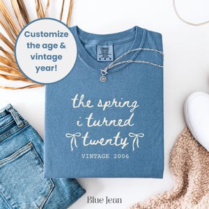 May include: A blue jean colored t-shirt with the text "the spring i turned twenty vintage 2006". A silver necklace with a pendant is on the shirt. The shirt is next to a pair of blue jeans and a white shoe.