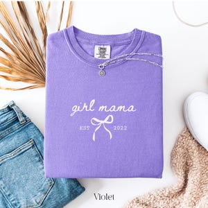 May include: A violet Comfort Colors crewneck sweatshirt with the text "girl mama" and a bow graphic. The text "EST 2022" is below the bow. A silver necklace with a pendant is draped on the shirt. A pair of blue jeans and a white shoe are in the background.