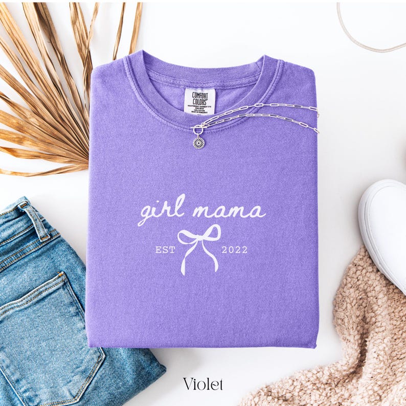 May include: A violet Comfort Colors crewneck sweatshirt with the text "girl mama" and a bow graphic. The text "EST 2022" is below the bow. A silver necklace with a pendant is draped on the shirt. A pair of blue jeans and a white shoe are in the background.