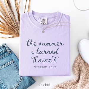 May include: A light purple short-sleeve t-shirt featuring the dark gray text "the summer i turned nine" with two small bow illustrations and the text "VINTAGE 2017" below. The shirt has a Comfort Colors brand label at the inner neckline.