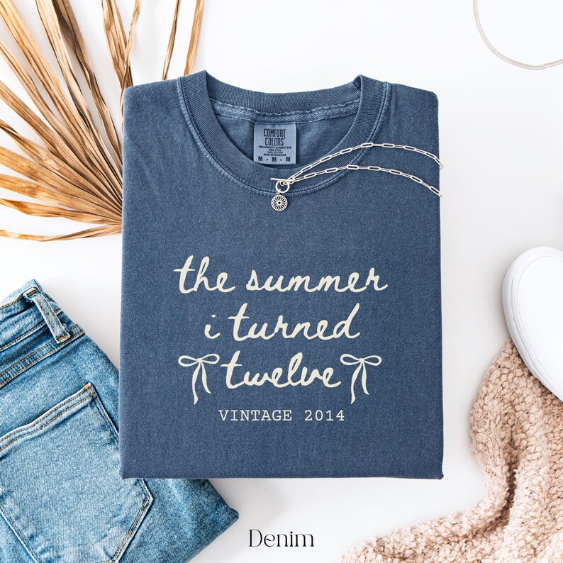 May include: A denim blue t-shirt with the text "the summer i turned Twelve Vintage 2014" in white script. A silver necklace with a sun pendant rests on the shirt. The word "Denim" is at the bottom.