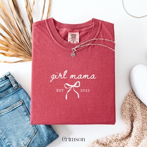 May include: Crimson-colored t-shirt with the words "girl mama" and a bow graphic, with "EST 2022" printed below. A silver necklace with a pendant rests on the shirt. The shirt is folded on a white surface, with blue jeans and a beige sweater visible.