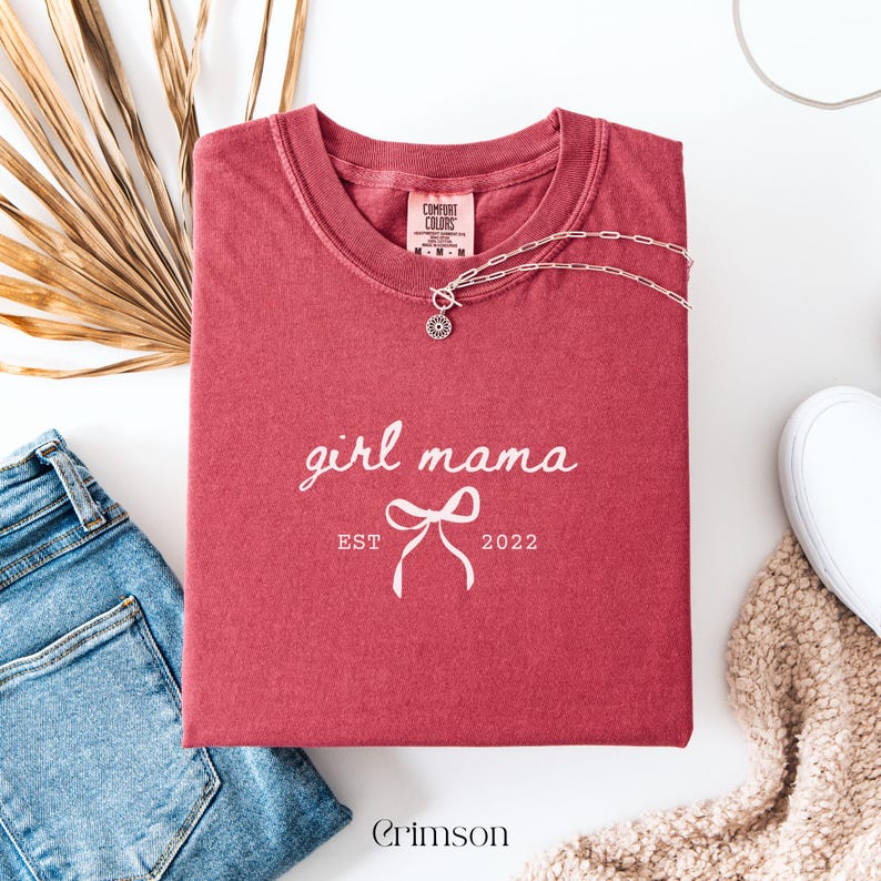 May include: Crimson-colored t-shirt with the words "girl mama" and a bow graphic, with "EST 2022" printed below. A silver necklace with a pendant rests on the shirt. The shirt is folded on a white surface, with blue jeans and a beige sweater visible.