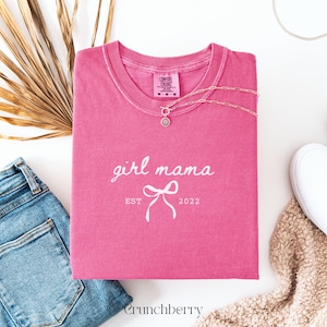 May include: A pink short-sleeve t-shirt with a crew neckline featuring the white text girl mama, a graphic of a bow, and the text EST 2022. The shirt has a Comfort Colors label at the inner neck and is displayed with a silver chain necklace.