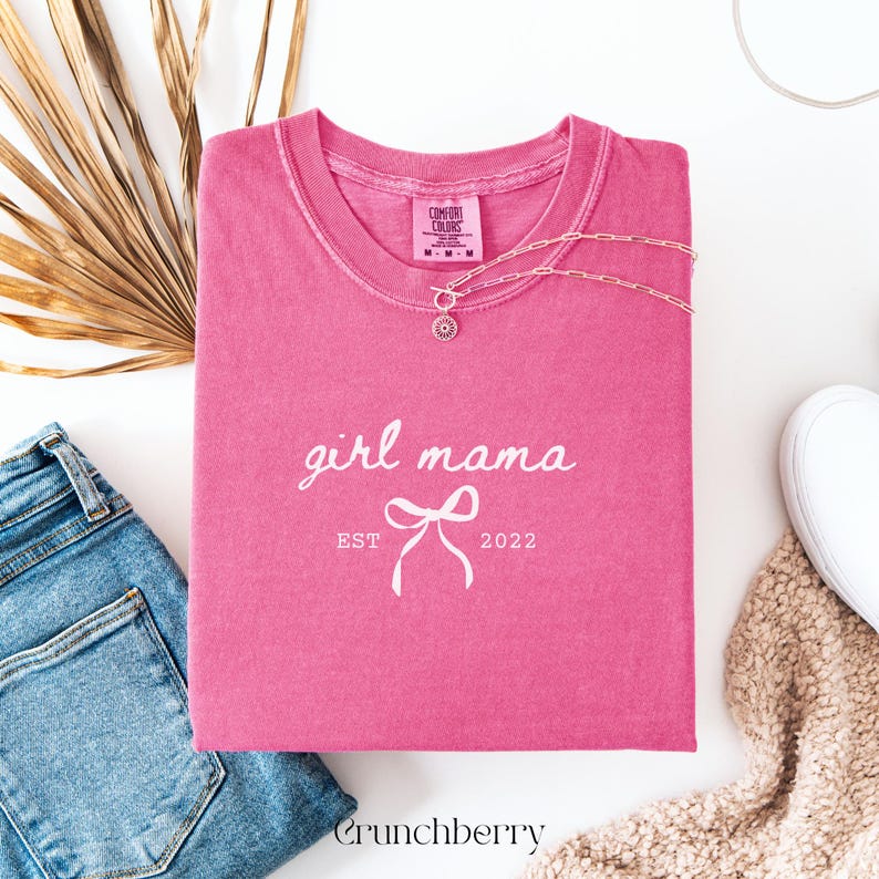 May include: A pink short-sleeve t-shirt with a crew neckline featuring the white text girl mama, a graphic of a bow, and the text EST 2022. The shirt has a Comfort Colors label at the inner neck and is displayed with a silver chain necklace.