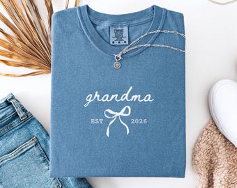 May include: A blue jean colored t-shirt with the word "grandma" in white script, a bow graphic, and the text "EST 2026". A silver necklace with a pendant is draped on the shirt. A pair of blue jeans and a white shoe are also visible.