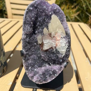 May include: A large amethyst geode with a dark purple exterior and a cluster of white and reddish-brown crystals in the center. The geode is mounted on a black metal stand, showcasing its natural beauty.