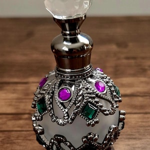 May include: An ornate perfume bottle with a clear, faceted stopper. The silver-toned metal bottle is adorned with purple and green gemstones. The bottle has a frosted glass body and intricate filigree detailing.