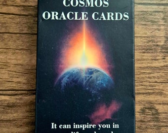 Op de afbeelding: Een zwarte doos met de tekst "COSMOS ORACLE CARDS" in wit. De doos heeft een kleurrijk beeld van de aarde met een helder oranje licht dat erop schijnt. De tekst "It can inspire you in your life and work." is in wit gedrukt aan de onderkant van de doos.