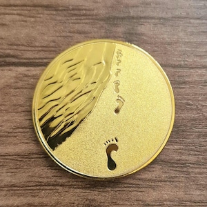 Könnte beinhalten: Eine runde goldfarbene Münze mit einem Design von Fußabdrücken am Strand. Die Münze hat einen erhöhten Rand und eine glatte Oberfläche.
