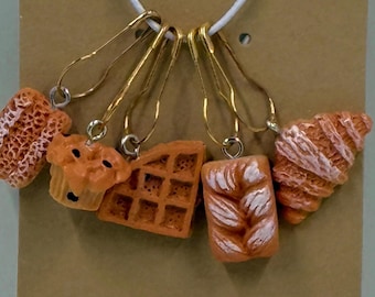 May include: A collection of orange-colored food-themed stitch markers. The set includes a waffle, croissant, muffin, and two other baked goods. Each charm is attached to a gold-colored safety pin and strung on a white cord.