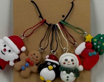May include: A collection of Christmas-themed charms, including Santa Claus, a gingerbread man, a penguin, a snowman, a stocking, and a Christmas tree. Each charm is brightly colored and attached to a black cord with a silver clasp. The charms are displayed on a brown card.