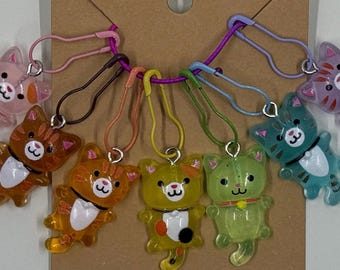 Puede incluir: Una colección de colgantes con forma de gato de colores con cierres de imperdible. Los colgantes son de varios colores, incluyendo naranja, amarillo, verde y rosa. Cada colgante tiene una cara pequeña y redonda y un diseño de dibujos animados.