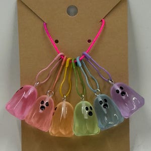 May include: A set of seven colorful ghost-shaped stitch markers hanging from a brown card. Each ghost is a different color, including pink, orange, yellow, green, blue, and purple. The ghosts have black dot eyes and are attached to the card with a pink cord.