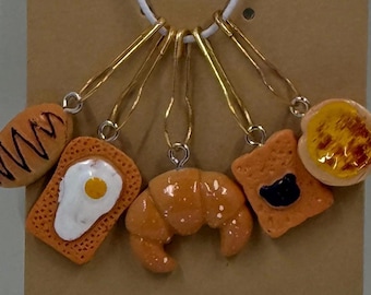 May include: A necklace featuring miniature food charms, including a croissant, toast with a black jelly, toast with an egg, and a bread roll. The charms are orange and brown, suspended from gold-colored pins and a white cord.