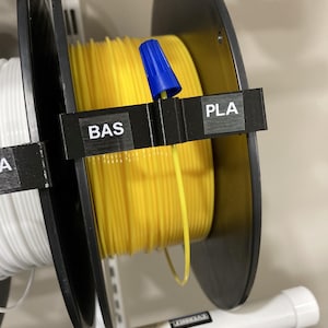 Può includere: Due bobine di filamento per una stampante 3D. La bobina a sinistra è bianca e la bobina a destra è gialla. Le bobine sono etichettate "PLA" e "BAS".