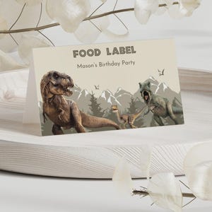 May include: A folded paper food label card featuring a beige background with the text FOOD LABEL and Mason's Birthday Party. The design includes illustrations of three dinosaurs in a mountain landscape with pine trees and small flying pterosaurs.