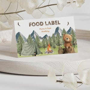 May include: A folded white card with the text FOOD LABEL and Mason's First Birthday. The illustration features a green tent, a campfire, pine trees, mountains, and a brown bear sitting on a tree stump in a forest setting.