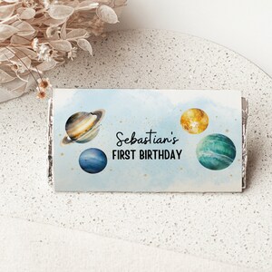 Puede incluir: Un envoltorio de chocolate azul y blanco con un diseño de acuarela de planetas y estrellas. El texto en el envoltorio dice "Sebastian's First Birthday".