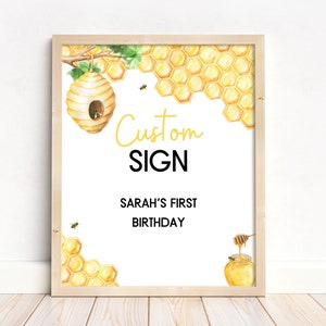 May include: A printable sign with a watercolor illustration of a honeycomb, a beehive, and a honey pot. The text on the sign reads "Custom Sign" and "Sarah's First Birthday".