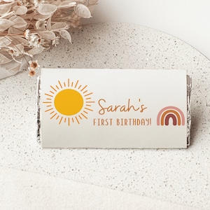 May include: A white chocolate bar wrapper with a yellow sun and a rainbow design. The text on the wrapper reads "Sarah's First Birthday!"