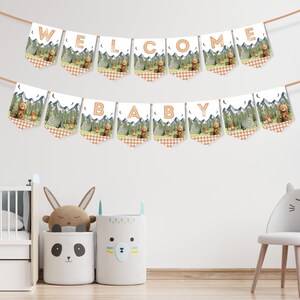 Op de afbeelding: Een witte banner met bruine en groene pennanten met een bosmotief waarop 'Welkom Baby' staat. De banner hangt aan een witte muur met een wieg en een stoel op de achtergrond.
