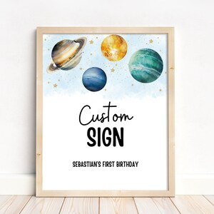 Puede incluir: Un cartel imprimible con un fondo de acuarela azul y estrellas doradas. El cartel dice "Custom Sign" y "Sebastian's First Birthday" con planetas de acuarela azul, verde y amarillo.