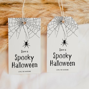 Puede incluir: Dos etiquetas de regalo blancas con diseños de telarañas negras y arañas negras colgando de las telarañas. El texto en las etiquetas dice "Have a Spooky Halloween" con "LOVE, THE WATSONS" en la parte inferior.