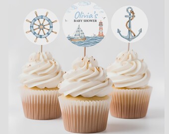 Puede incluir: Tres cupcakes coronados con círculos de papel de temática náutica en palillos de madera. El círculo izquierdo muestra un timón azul, el central un velero, un faro y el texto Olivia's BABY SHOWER, y el derecho muestra un ancla gris.
