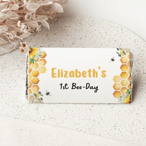 May include: A white chocolate bar wrapper with a yellow honeycomb pattern and a sunflower design. The wrapper says "Elizabeth's 1st Bee-Day".